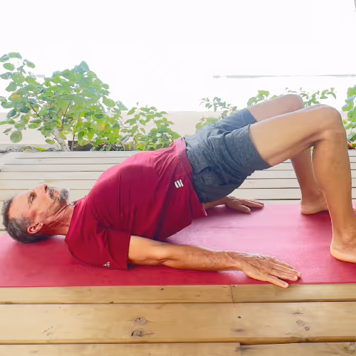 Mann macht eine Brücke-Übung auf einer Yogamatte im Freien mit Pflanzen im Hintergrund.