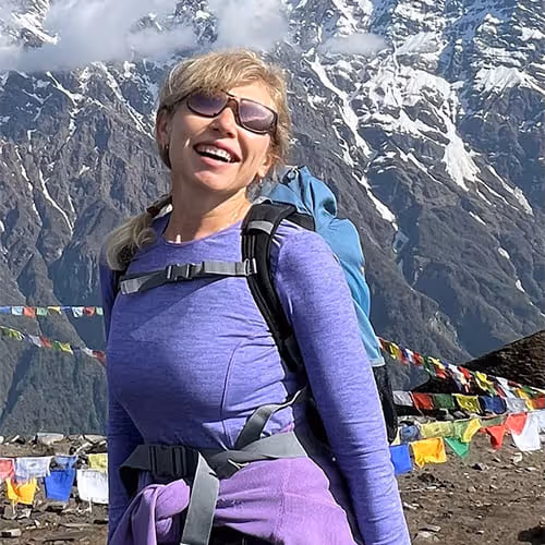 Frau mit Sonnenbrille und Rucksack, lachend vor schneebedeckten Bergen und bunten Gebetsfahnen.