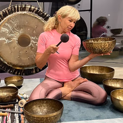 Frau im Yoga-Sitz mit Klangschalen in einem Meditationsraum, hält eine Schale und einen Schlägel.