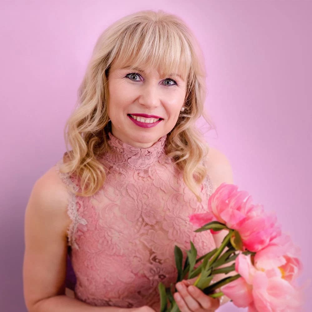 Frau mit blonden, lockigen Haaren in einem rosa Spitzenkleid hält einen Strauss pinker Blumen vor einem rosa Hintergrund das ist Veronika Mutti - Expertin für Massage und Coaching in Schweiz