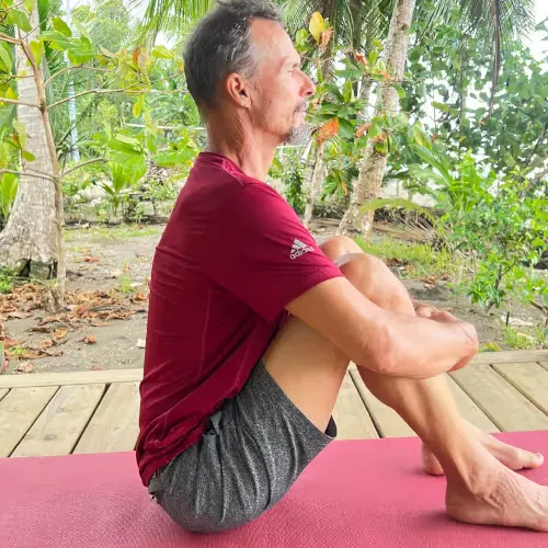Mann sitzt auf Yogamatte im Schneidersitz und meditiert im Freien unter Bäumen.