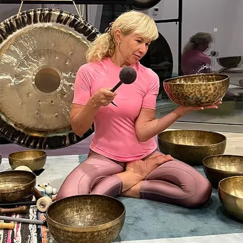 Frau im Yoga-Sitz mit Klangschalen in einem Meditationsraum, hält eine Schale und einen Schlägel.