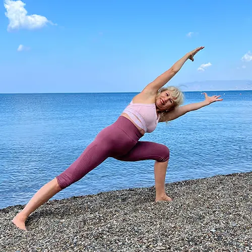 Frau macht eine Yoga-Dehnstellung am kiesigen Strand vor einem ruhigen See unter blauem Himmel.
