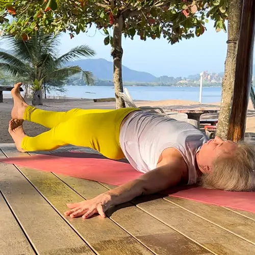 Frau in gelber Leggings macht Yoga-Übung seitlich liegend auf einer Holzterrasse mit Blick auf Strand und Meer.